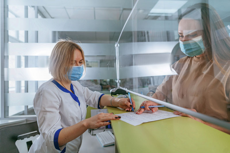 Female nurse explaining patient how to fill form at hospital reception. Doctor and patient wearing protective face masks and using protective glass screen.