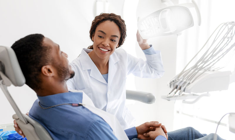 Male patient sitting in dental office, female dentist turning on a lamp before treatment, smiling to male patient.