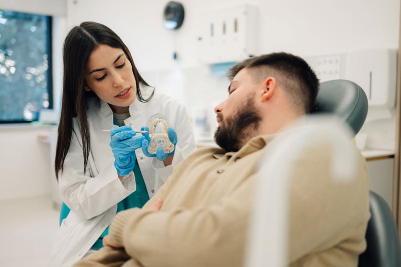 Female dentist wearing blue gloves and pointing at a teeth model while explaining something to male patient sitting in dentist chair during