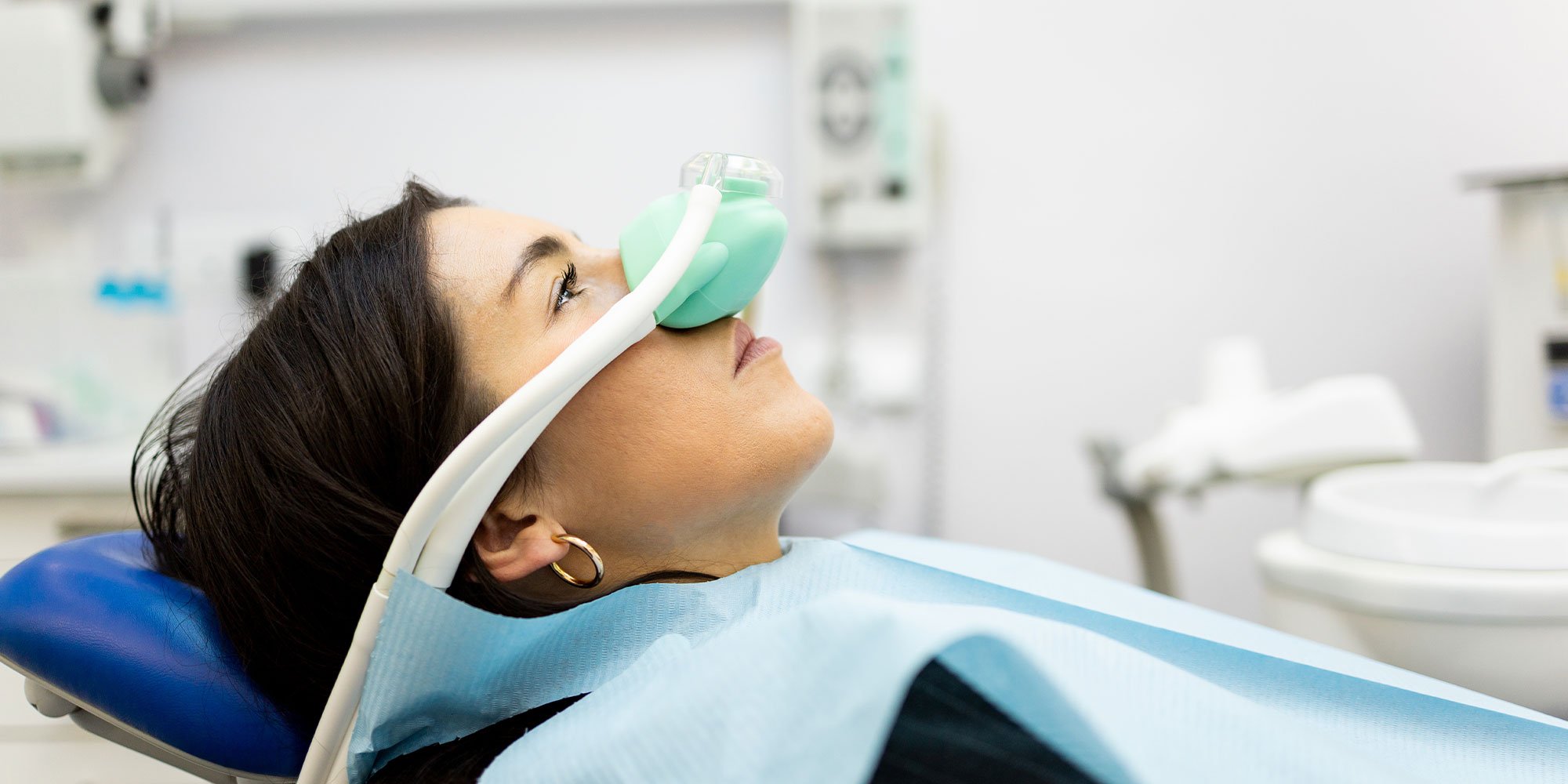 An adult woman sits in a dentist's office wearing a nasal mask to inhale nitrous oxide